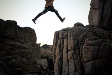 person jumping on big rock under gray and white sky during daytime